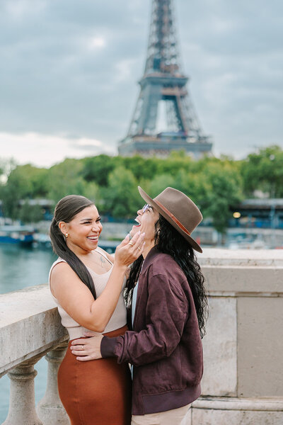 Lesbian couple laughing and embracing after a proposal near the Eiffel Tower at Bir-Hakeim Bridge during a romantic Paris photoshoot.