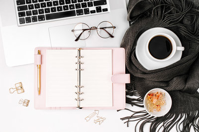 Stock image of pink planner, coffee in a white mug, glasses and laptop with a gray scarf