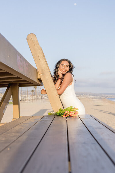 A woman with long brown hair, wearing a long white dress standing on a wooden latter on a lifeguard statue, leaning on the ladder and looking to her left at the camera with a bouquet of red and orange flowers resting below her on the wooden step in Newport Beach.