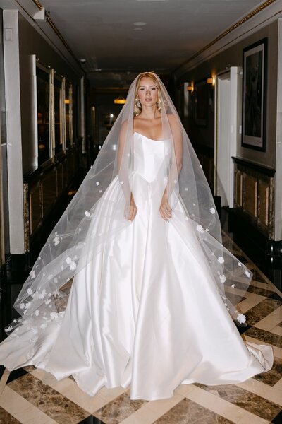 Bride in a satin gown with long train and floral veil, standing in a hallway.