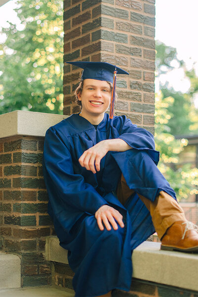 Senior guy wearing cap and gown and leaning against a brick wall