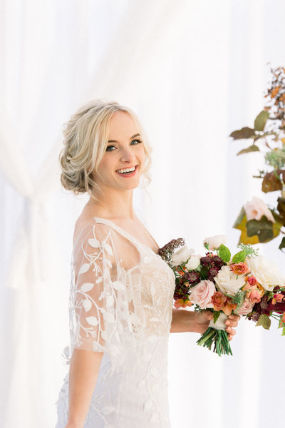 Bride with textured low bun and natural, clean makeup smiles while holding flowers, Indianapolis Wedding Hair and Makeup