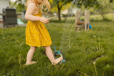 toddler girl places foot on sprinkler while wearing a cute yellow dress