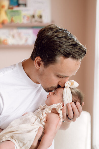 A father gently kisses his newborn’s forehead during a heartfelt in-home family photography session in Northern Virginia.