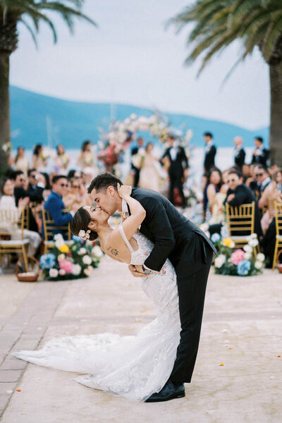Groom kissing bride during seaside ceremony surrounded by guests and floral decor, photographed by wedding photographer in Toronto