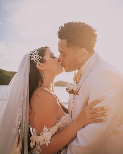 A bride and groom share a passionate kiss during their sunset beach wedding, with golden light enhancing their special moment.