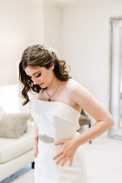 A bride with wavy brown hair and a hairpiece looks down as she adjusts her strapless white wedding dress, standing in a softly lit room with a sofa and a large mirror in the background.