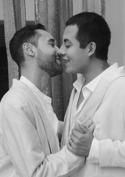 Partners in white suits holding hands, reaching for a kiss – LGBTQ wedding captured beautifully.