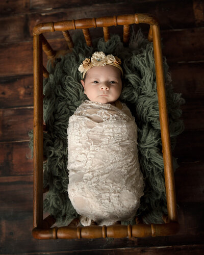 Newborn girl wrapped in a white lace wrap laying in a crib.