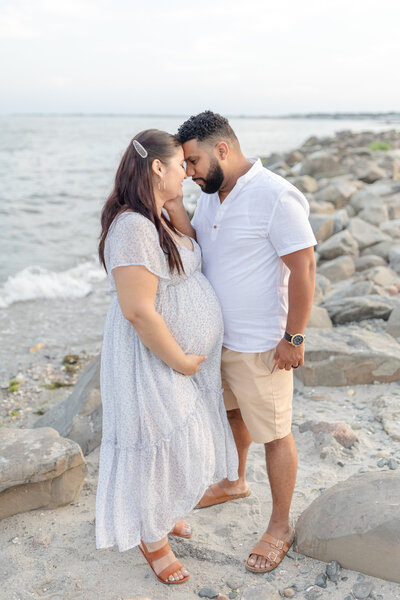 A husband and his pregnant wife stand on the beach during golden hour, their foreheads gently touching as they share a quiet, intimate moment, filled with love and anticipation for their baby’s arrival.