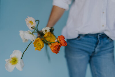 Close up photo of gathered flowers in orange, yellow and cream being held to the side by Alexandra in front of a blue background