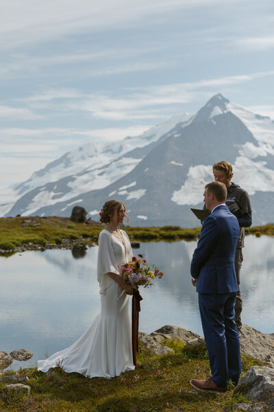 Elopement couple says vows with officiant by Alaska mountain lake.