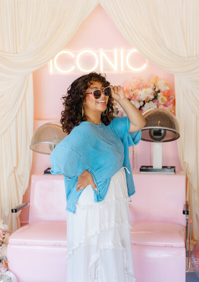 Joyful woman in sunglasses and flowy skirt posing in front of a retro salon backdrop with pink chairs and florals — styled portrait session by Greenville South Carolina portrait photographer Dani Lacey Photographs.