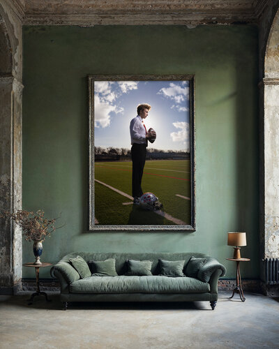 A high school senior portrait of a young man in a white shirt and red tie, holding a football on a field. The photo is framed in a vintage, weathered green frame displayed on a wall in a rustic room with a vintage green sofa, a tall lamp, and a side table with a flower vase.