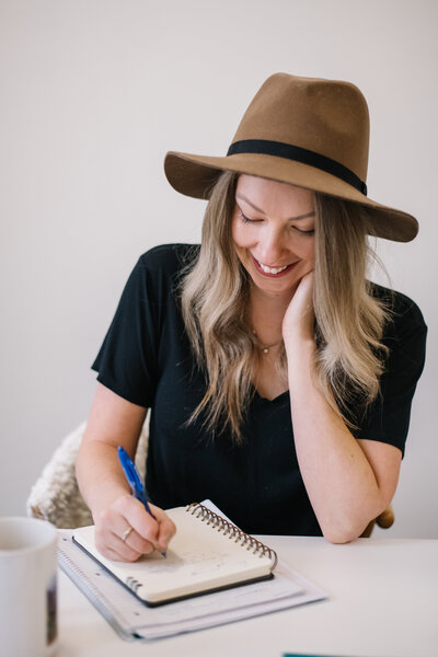 Sammy wearing a black shirt and brown hat smiling while writing in a notebook with a blue pen