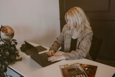 A blonde woman is sitting at a white desk, looking down at her laptop, typing.