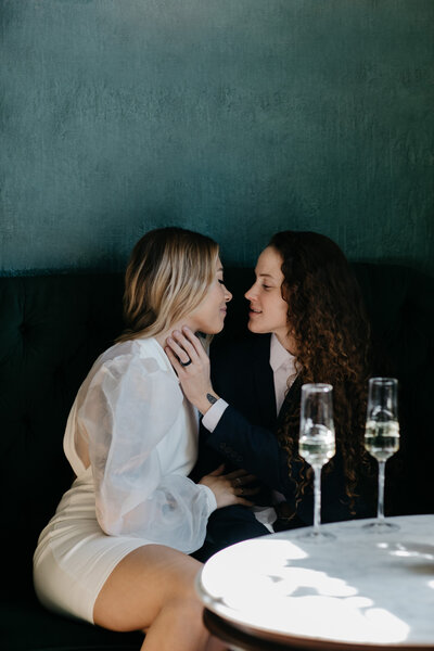 Female couple sitting down with champagne share a mid-kiss moment