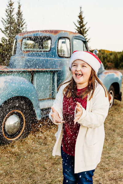 adorable young girl wearing a santa hat and playing with the snow in front of a blue truck for her Christmas photo session in Wild Rose WI