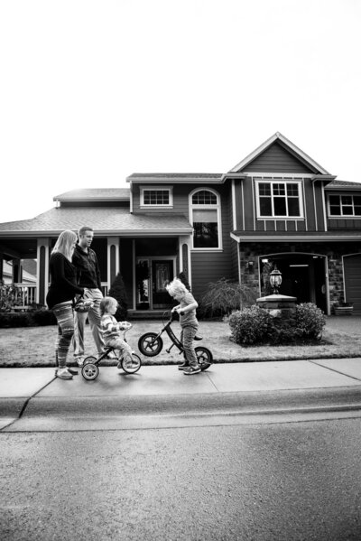 Family walking on sidewalk in front of their home with boys riding tricycles documentary scene black and white