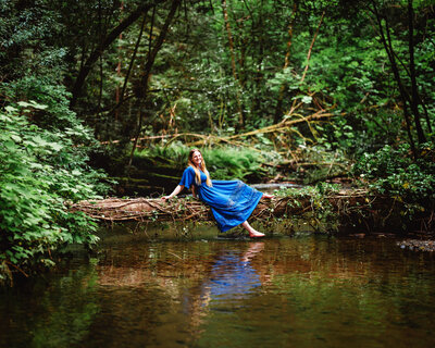 A senior girl wearing a blue gown sitting on a fallen log in a stream.