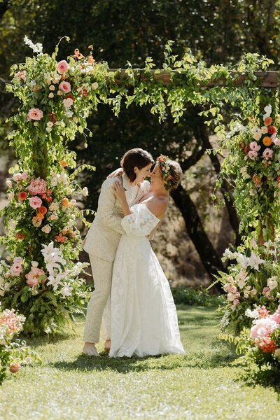 An LGBTQ couple celebrates at their wedding ceremony under colorful flower arrangements.