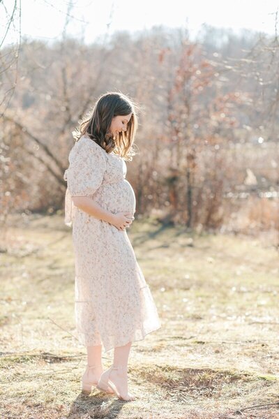 A woman stands outside in a floral dress holding her pregnant belly and gazing down at it while being photographed by New Jersey Maternity Photographer Kate Voda Photography