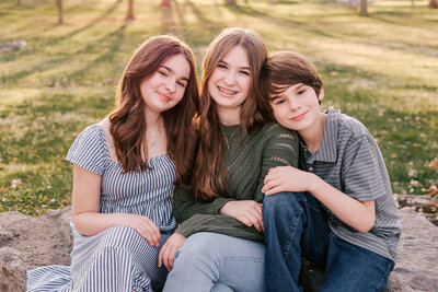 3 kids are sitting together outdoors with a sunset behind them. they are cozy and leaning on each other posing for their family photos.