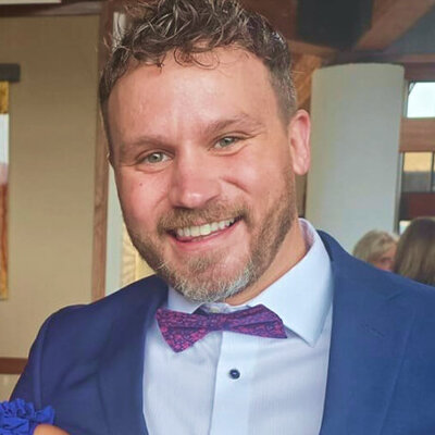 Man smiling, wearing a blue suit and purple tie while indoors. He has short blonde hair and a scruffy beard.