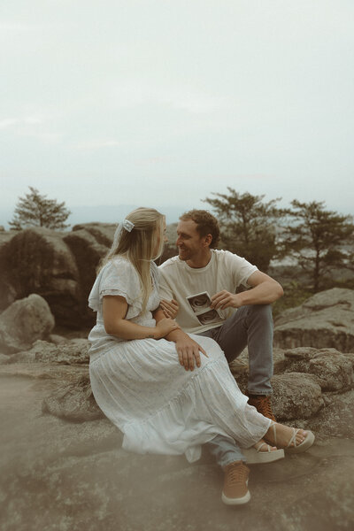 A couple sitting on rocks outdoors, sharing a moment while looking at ultrasound photo, surrounded by trees and a cloudy sky.