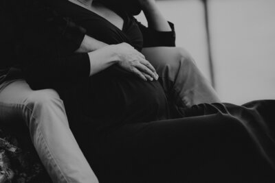 black and white image of the lower half of a pregnant women sitting in  between a man's legs with both their right hands resting on her stomach