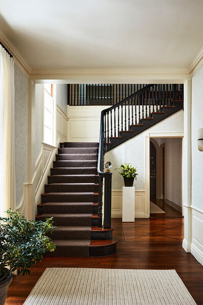 Interior decorators in Boston created a modern look in a historic Concord estate foyer with a black grand stair, cream walls, and geometric wallpaper blending old and new.