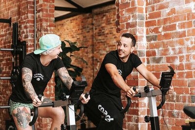 Friendly Interaction on Spin Bikes: Two gym-goers chatting and smiling while cycling on spin bikes in a Melbourne gym, emphasizing a fun and encouraging fitness environment.