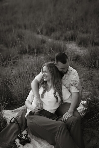 Black and white portrait of Stephen and Kaity sitting together and laughing in a field, captured for the About page of Rainstorm Photo & Video. Husband and wife wedding photographers and videographers based in Indiana.