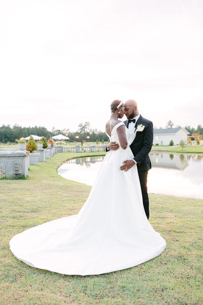 A bride in a stunning off-the-shoulder gown shares an intimate embrace with her groom by the lake at Chateau 1800 in Savannah. A timeless and elegant wedding moment captured at golden hour by Amia Marcell Photography.