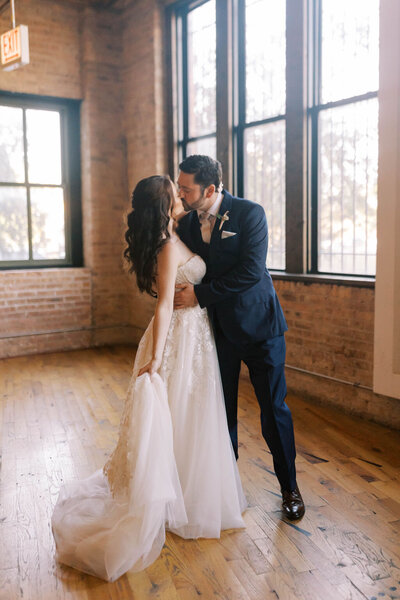Groom in blue tux holds bride in flowy gown around the waist as they share a kiss at the wedding venue