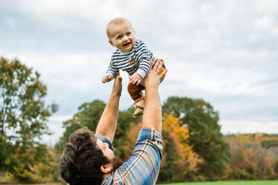 Father throwing baby son in the air with fall foliage in the background
