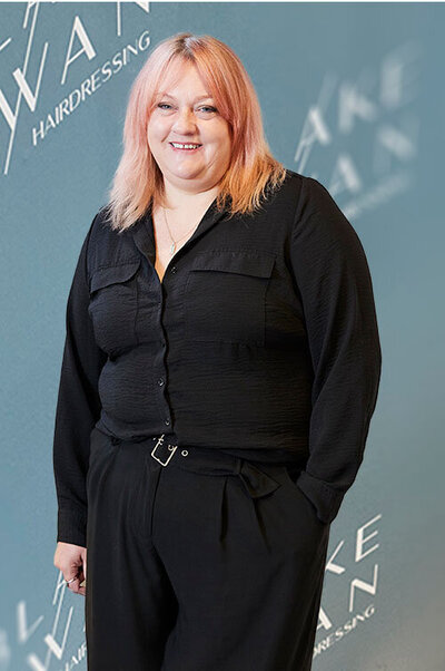 Owner & Hairdresser, Louise with pastel pink hair in black attire, standing against a branded Blake & Kwan Hairdressing backdrop.