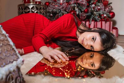 Mother in red dress smiling beside Christmas tree mirroring her face in Calini Weddings Studio – elegant festive portrait with wrapped gifts and golden ornaments.