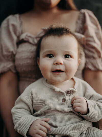 A baby smiles at the camera while wearing, and it will be a colored thermal shirt as his mother holds him during a sunset photography session.