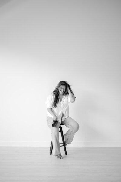 A black and white portrait photo of Hiba in a white studio. She's sitting on a stool and candidly smiling at the ground, camera in hand.