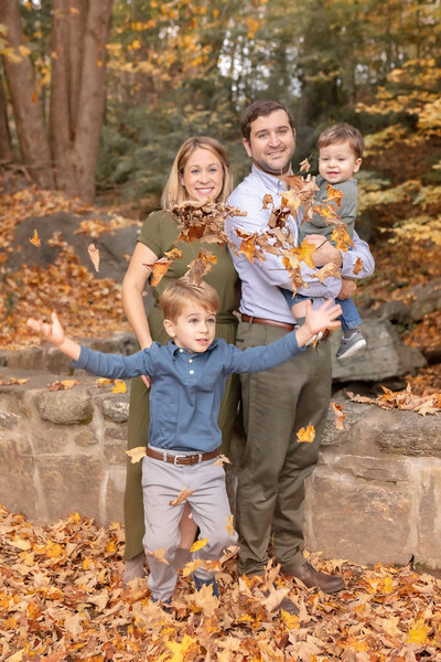 happy family of four in an autumn setting with young son throwing fall leaves in the air surrounding them in fall colors