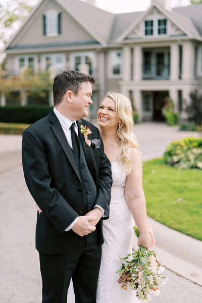 Groom looking at smiling bride holding bouquet in front of wedding venue in Oakville, Ontario