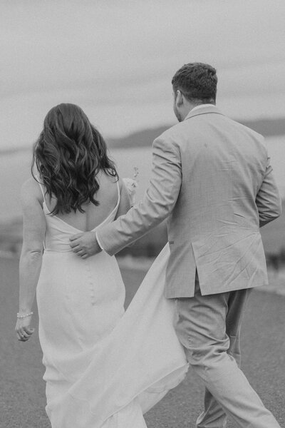 groom holding bride's train while walking in black and white