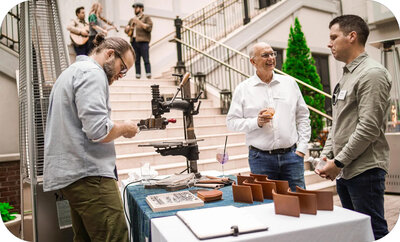 Person crafting leather goods at a small setup table while two others stand nearby talking in an indoor courtyard.