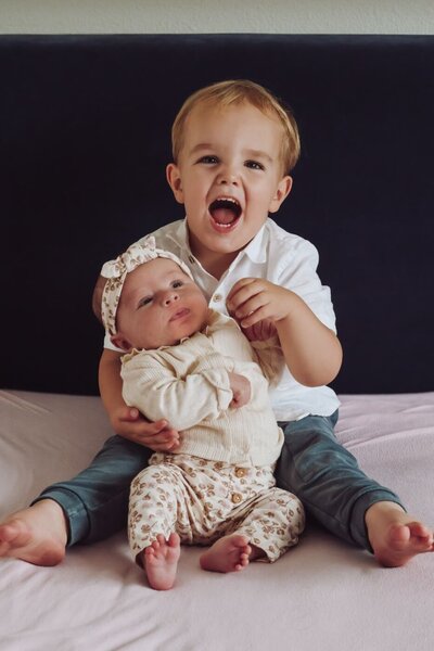 Een broer en zus poseren samen op een bed, waarbij de oudere broer enthousiast lacht en zijn zusje vasthoudt.