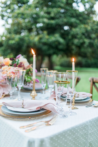 a wedding reception table with romantic taper candles and a large floral centerpiece. The table is set with a modern romantic table setting consisting of gold rimmed glassware, a gold glass charger, scalloped plates, and a blush cotton napkin. There is a white swiss dot linen on the table