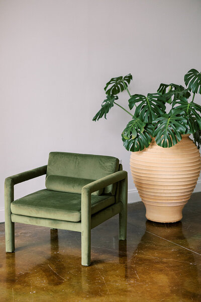 A green velvet chair with wooden armrests is positioned on a polished brown floor. Next to it is a large beige, textured vase containing lush green monstera leaves. The background is a plain light-colored wall.