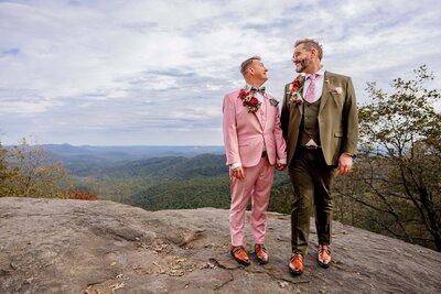 Two grooms stand side by side holding hands, looking at each other and smiling after eloping. The are standing on a large rock at the top of a mountain overlook. 