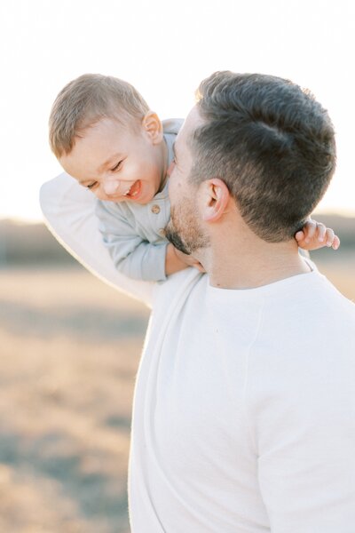 Hispanic dad holding toddler son over his shoulder laughing in a field during golden hour