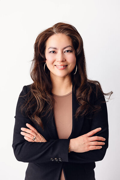 Woman with black jacket folds arms and smiles in a white studio.